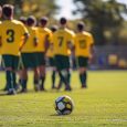 Un ballon de football sur la pelouse avec des jeunes joueurs flous en tenue jaune et vert en arrière-plan sur un terrain d’entrainement.